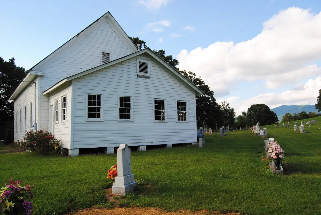 A white clapboard church with a gabled roof beside a grassy cemetery of headstones decorated with flowers, with green trees and a blue mountain ridge in the distance.