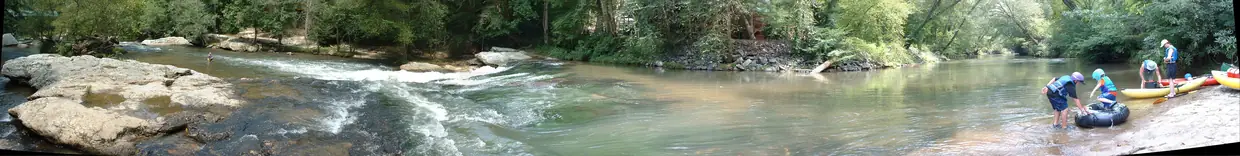 A wide panorama of a shallow rocky river with small rapids on the left calming toward the right, boulders and rhododendron along the banks and wooded hills behind.