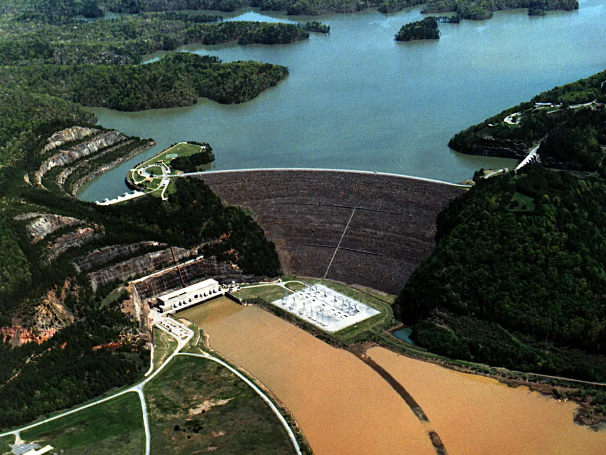 An aerial view of a large curved rock-fill dam holding back a wide lake with wooded islands, with a powerhouse, an electrical switchyard and a muddy brown river channel below the dam.