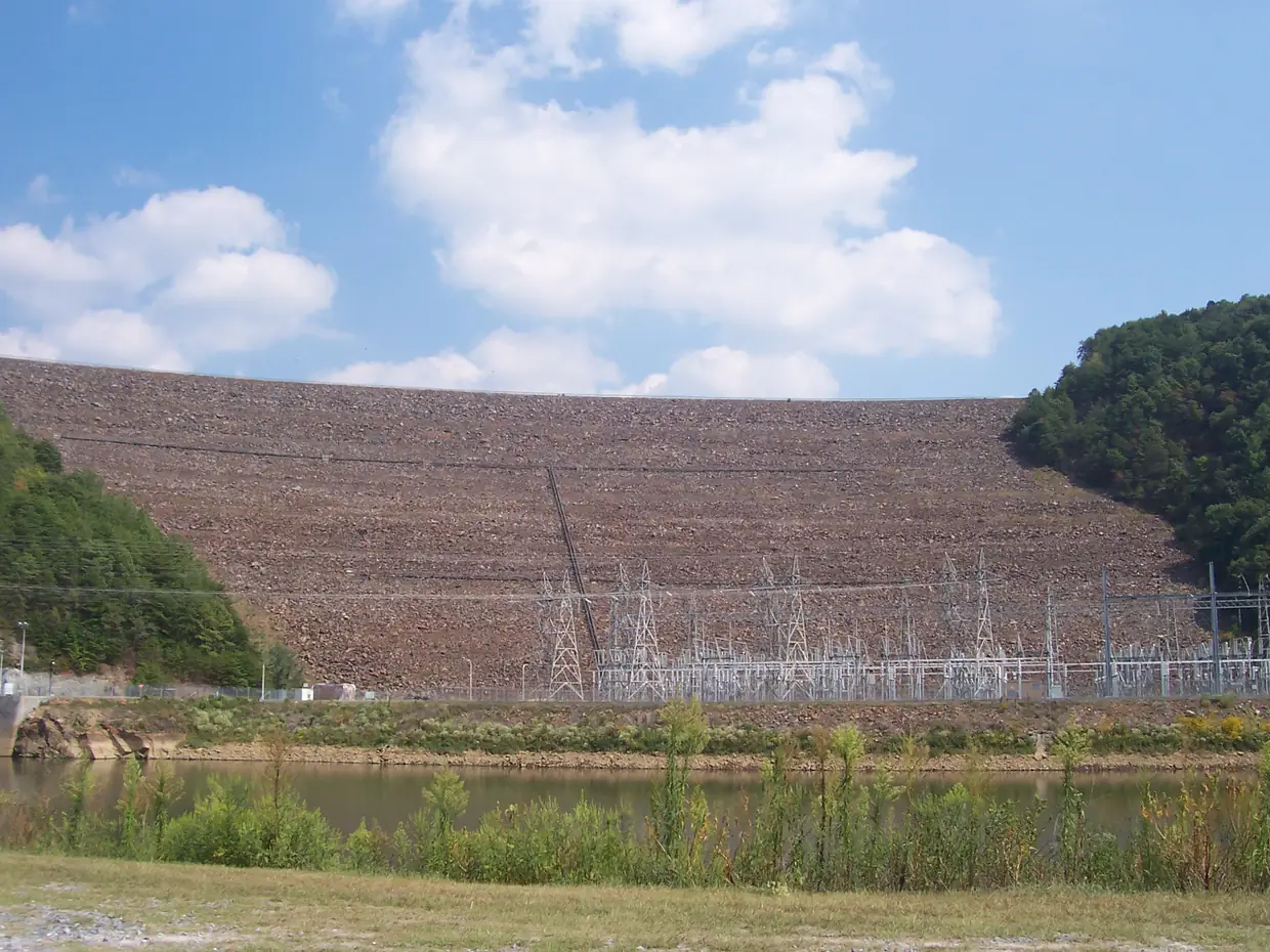 The broad sloping rock face of a large dam rising behind an electrical switchyard and a strip of water, with wooded hills at both sides and white clouds in a blue sky.