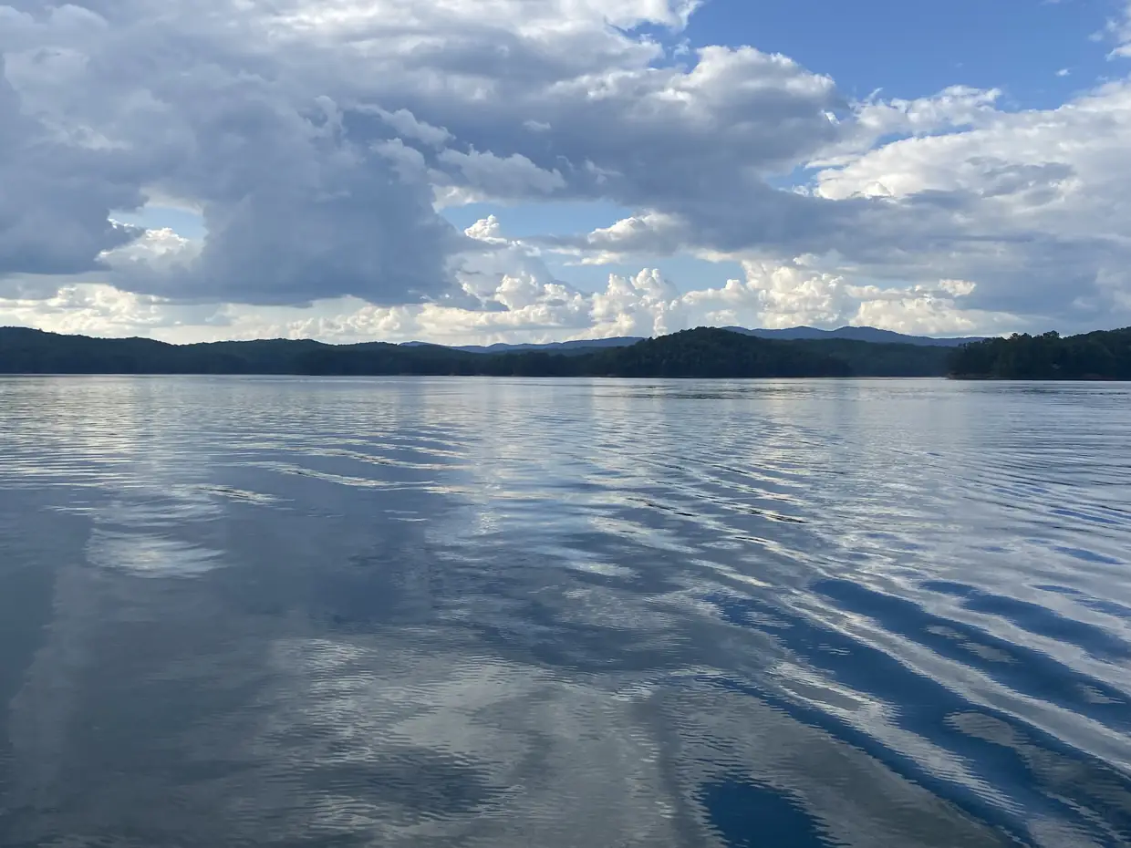 A calm, wide lake reflecting large white and gray clouds, with a dark wooded shoreline and low mountains along the horizon.