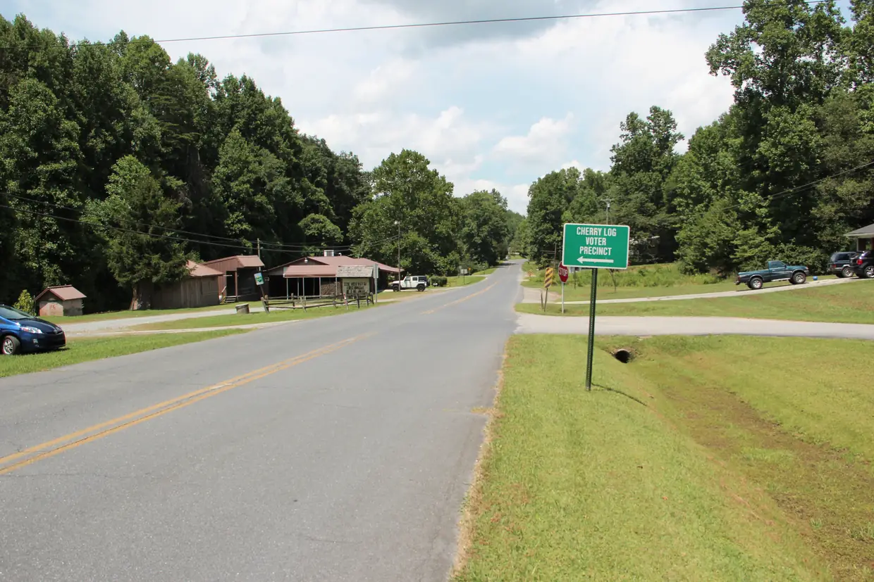 A two-lane road through a small rural community, with a green roadside sign reading CHERRY LOG VOTER PRECINCT, a few low buildings, parked vehicles and dense green trees.