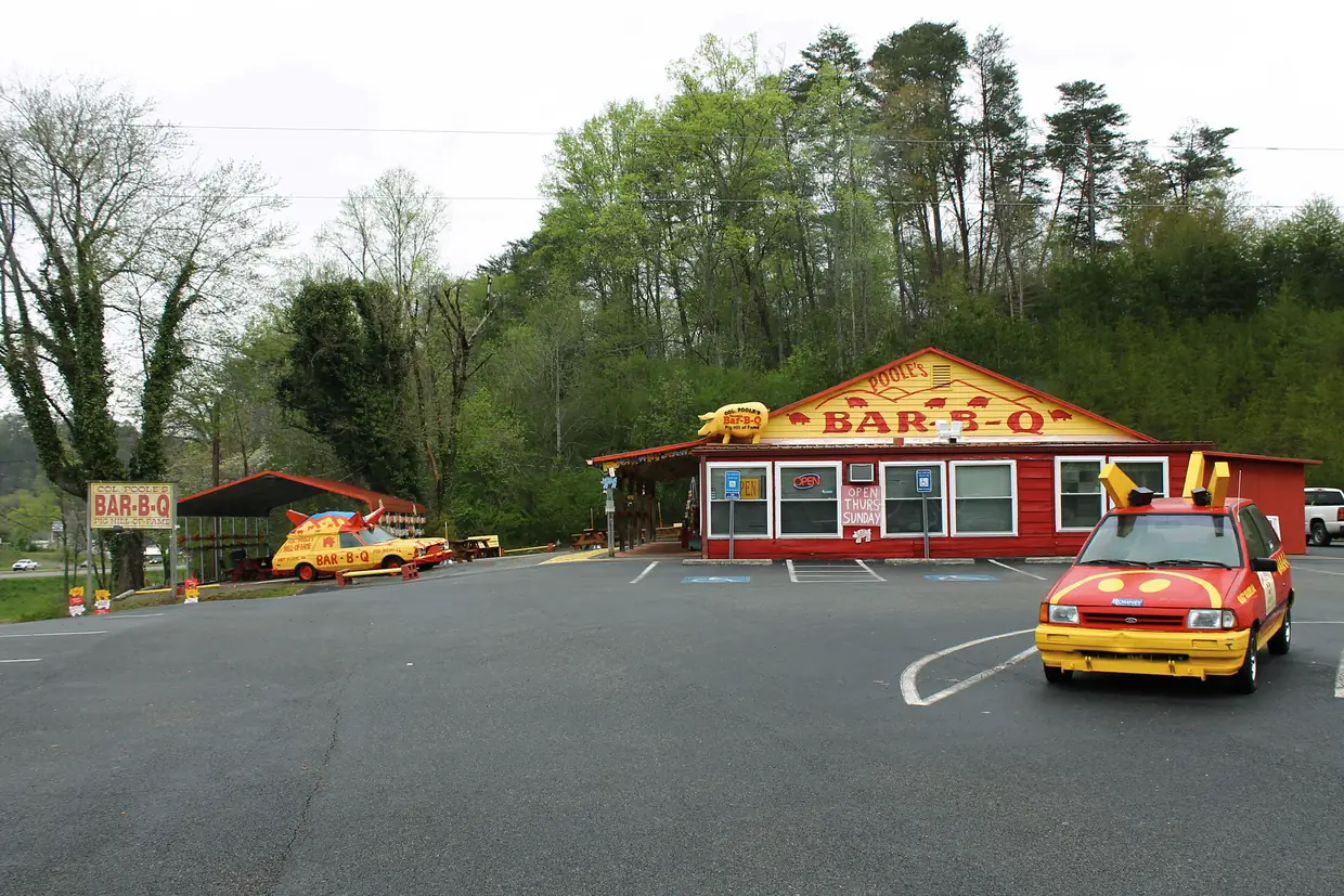 A red roadside barbecue restaurant with a yellow-trimmed gable sign reading BAR-B-Q, two yellow cars decorated as pigs in the parking lot and green wooded hills behind.