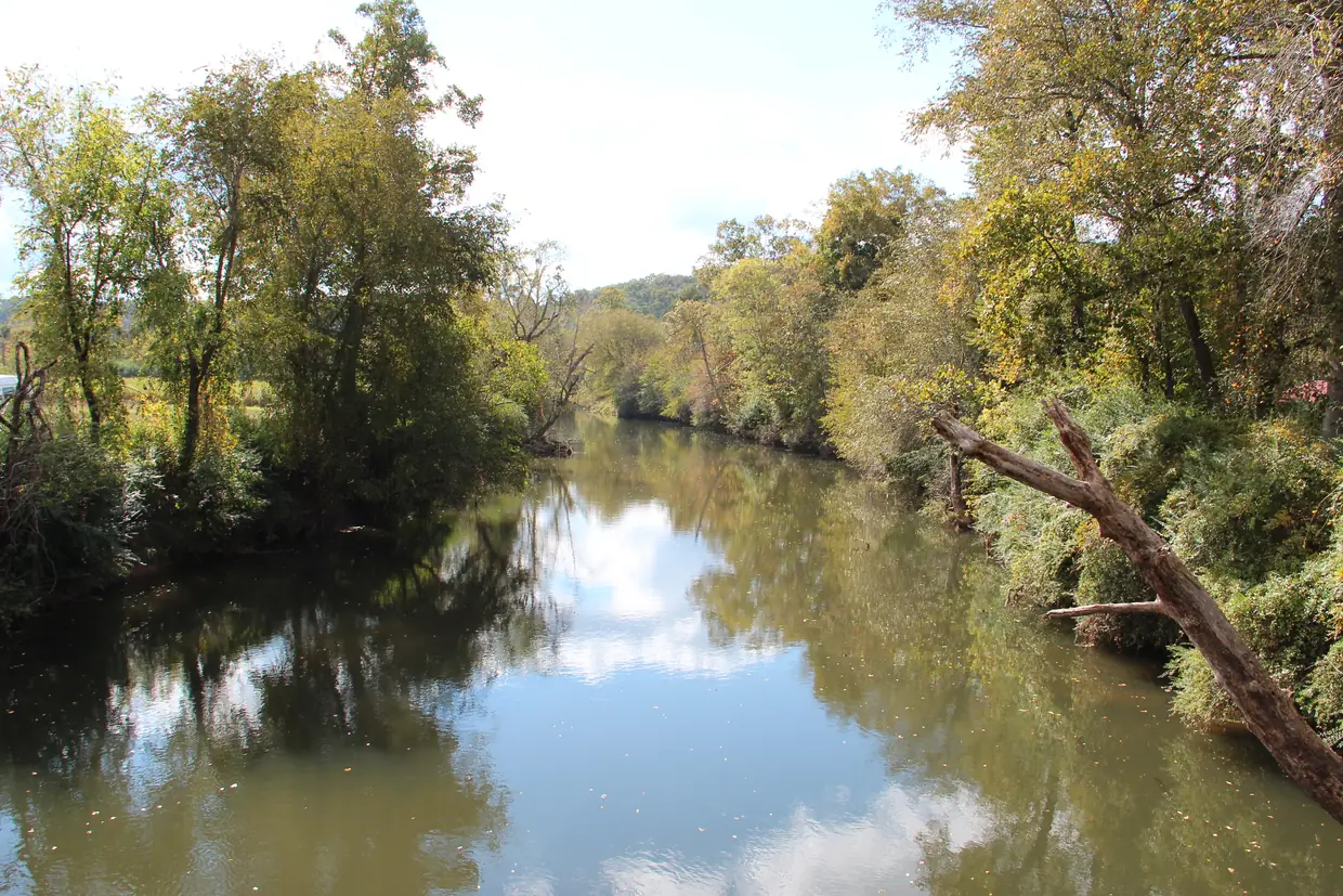 A calm green-brown river in a wooded valley seen from a bridge, with the sky and overhanging trees reflected in the water.