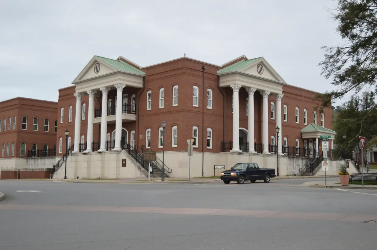 A red-brick courthouse with white columns and a clock tower seen from street level across a quiet two-lane road; a low white stone foundation, small trees at the edges and a clear sky.