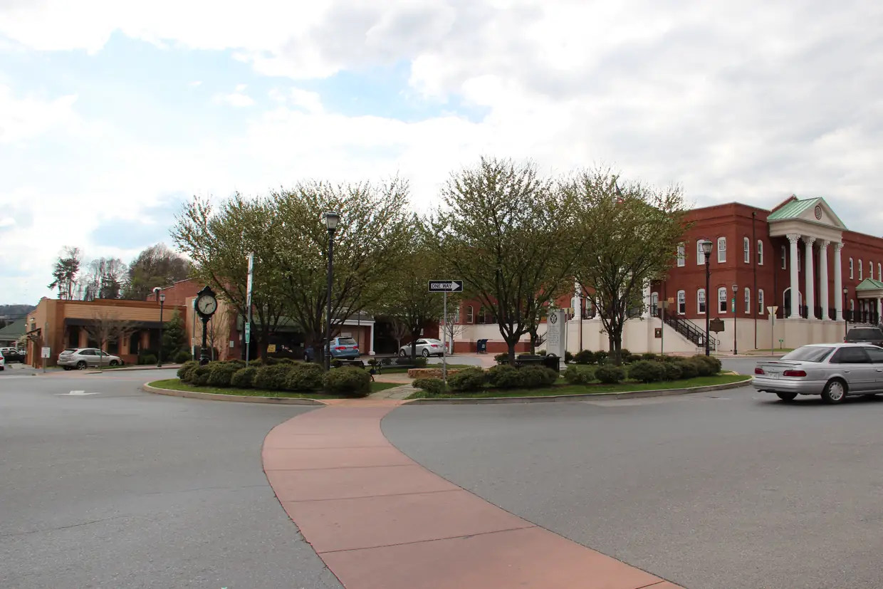 A landscaped roundabout with a brick-paved crosswalk, young leafing trees, a black post clock and street lamps in a small-town square; low brick storefronts on the left and a red-brick building with a white-columned portico on the right, under a partly cloudy sky.