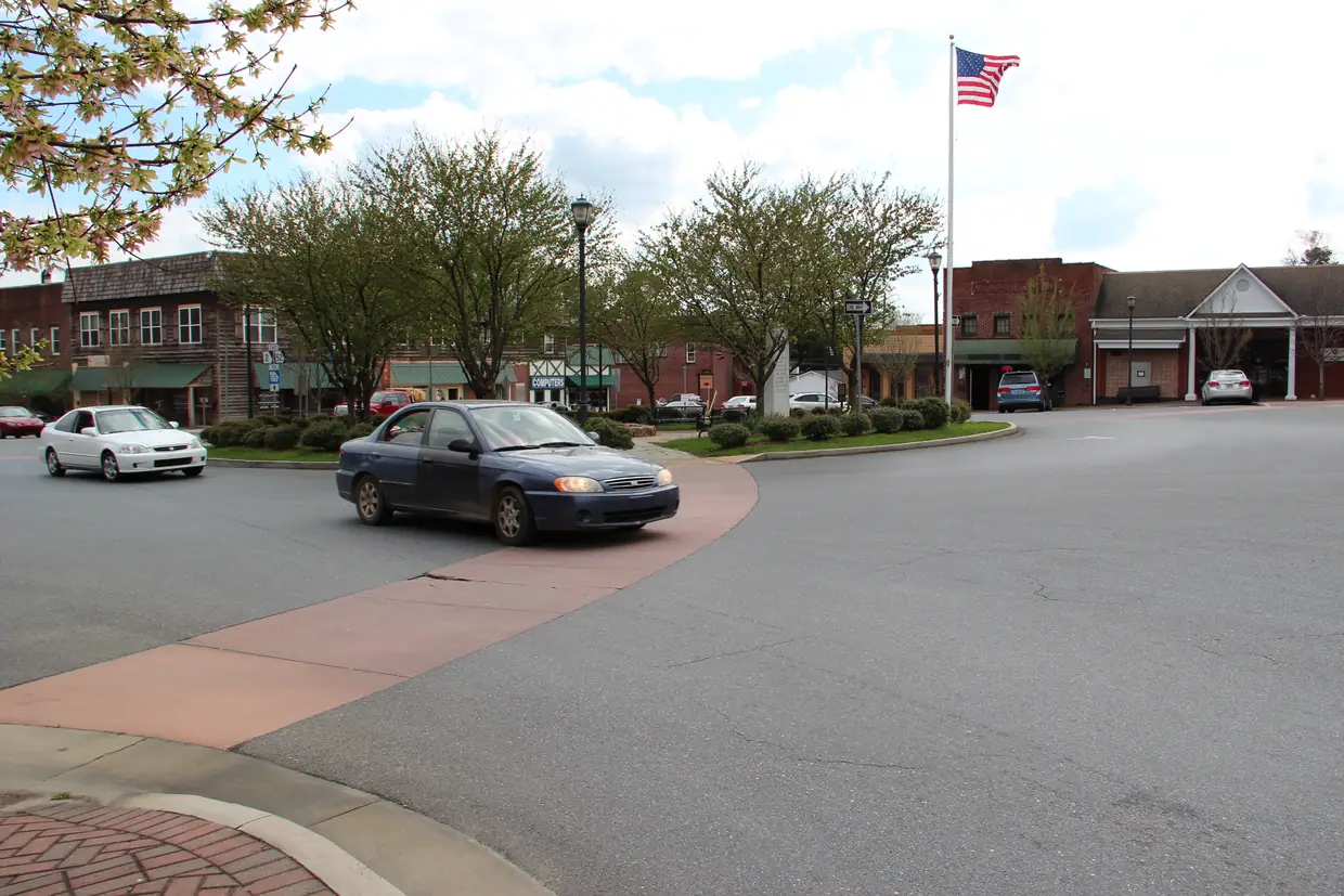 A small-town square seen across a curving street: a planted roundabout island with trees and a flagpole flying the U.S. flag, a dark sedan rounding the curve and a row of two-story brick storefronts with awnings behind.