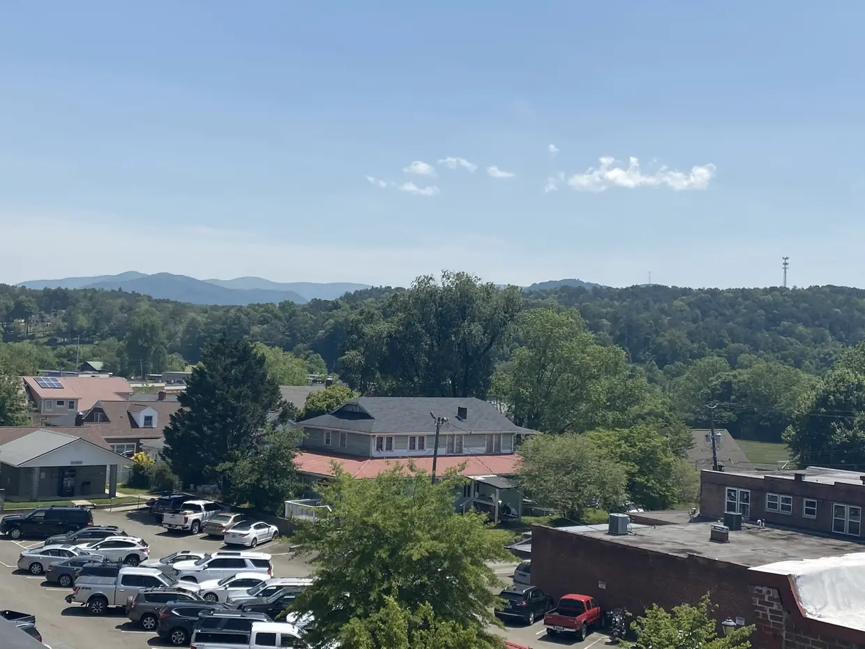 An elevated view over the low brick commercial buildings of a small town, a parking lot with cars in the foreground and wooded Blue Ridge foothills under a clear sky beyond.