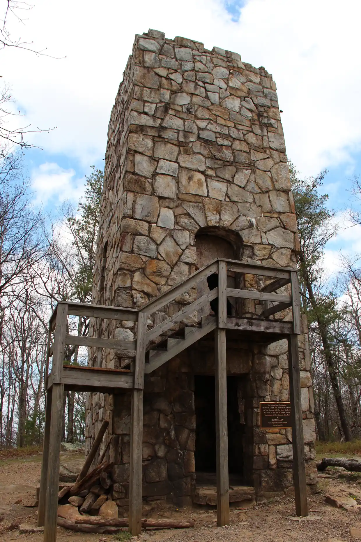 A tall rough-stone tower with an arched opening and a wooden stair landing at its base, standing among bare trees under a partly cloudy sky.