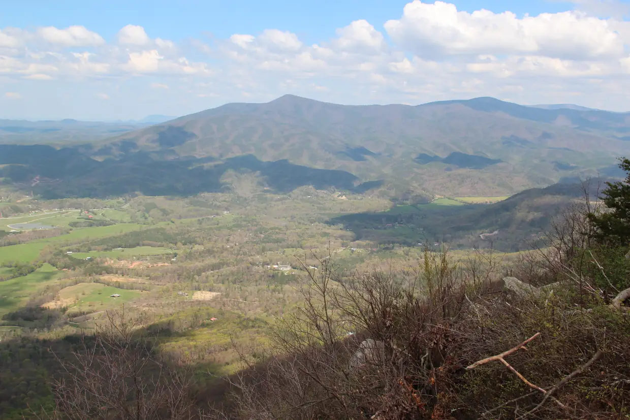 A high view over a broad green valley of farm fields and woods, with a rounded mountain range across the valley under white clouds and dry brush in the foreground.