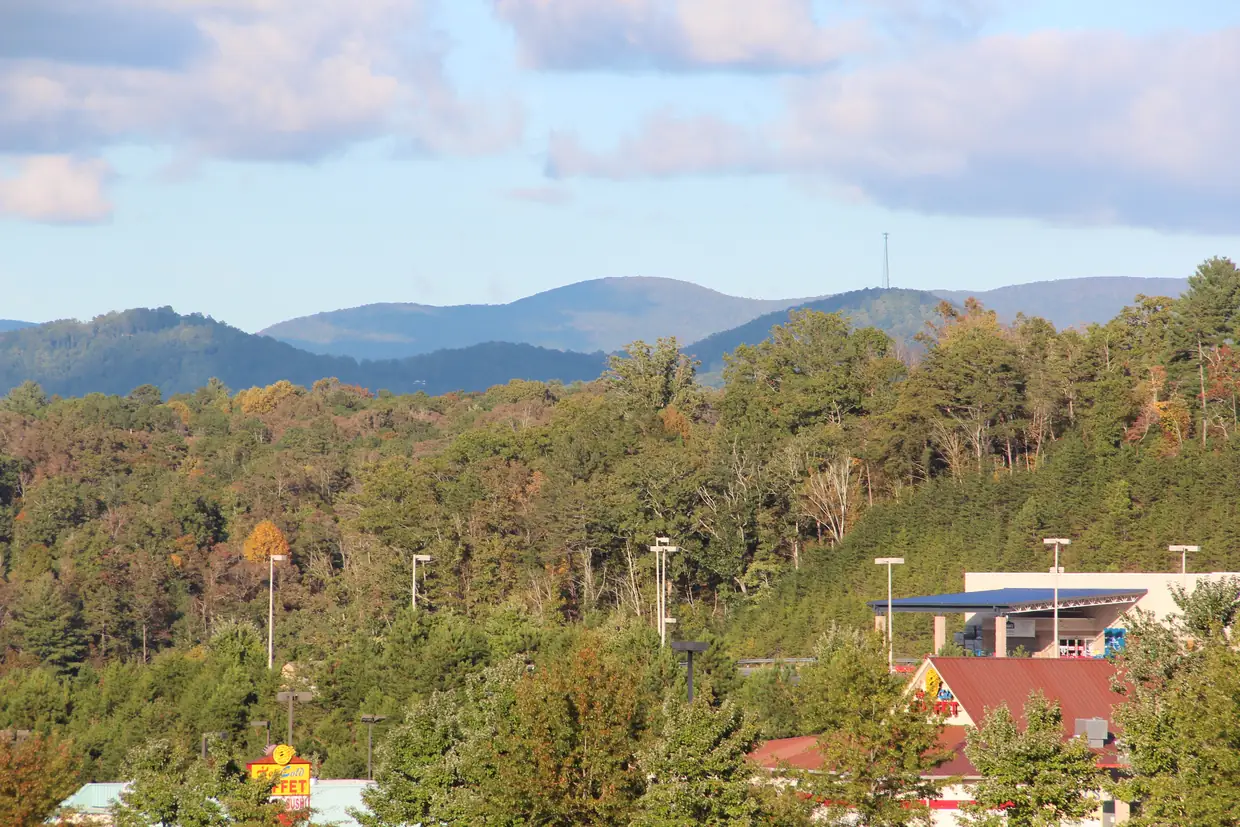 Wooded hills in early autumn color with rounded blue mountains and a radio tower on the skyline; light poles and the roofs of a few commercial buildings at the bottom edge.